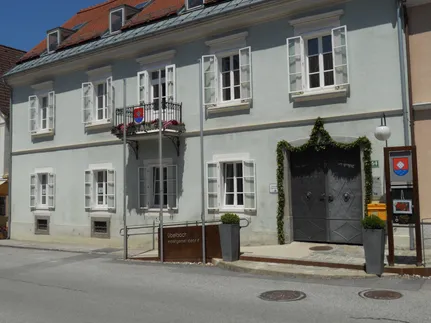 Ein Blick von der Straße auf ein zweistöckiges Gebäude mit rotem Dach und weißen Außenwänden, mit mehreren Fenstern, einem Balkon mit Blumen und einem großen Doppeltoreingang.
