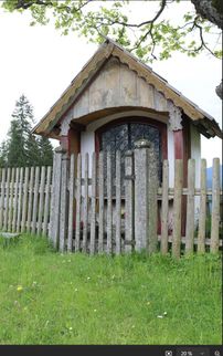 Ein kleines Holzhaus mit einem spitzen Dach und einem schwarzen Eisentor vor einem Holzzaun.