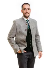 Ein lächelnder Mann in traditioneller deutscher Kleidung posiert für ein Foto. Er trägt eine graue Jacke mit Knöpfen, eine grüne Krawatte und eine Weste mit silbernen Knöpfen. Er hat die Hände in den Taschen.