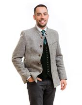 Ein lächelnder Mann in traditioneller deutscher Kleidung posiert für ein Foto. Er trägt eine graue Jacke mit Knöpfen, eine grüne Krawatte und eine Weste mit silbernen Knöpfen. Er hat die Hände in den Taschen.