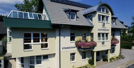 Ein Gebäude mit einem grauen Dach, Solarpanelen und einem Balkon mit rosa Blumen. Das Schild lautet 'STANDESAMT.'