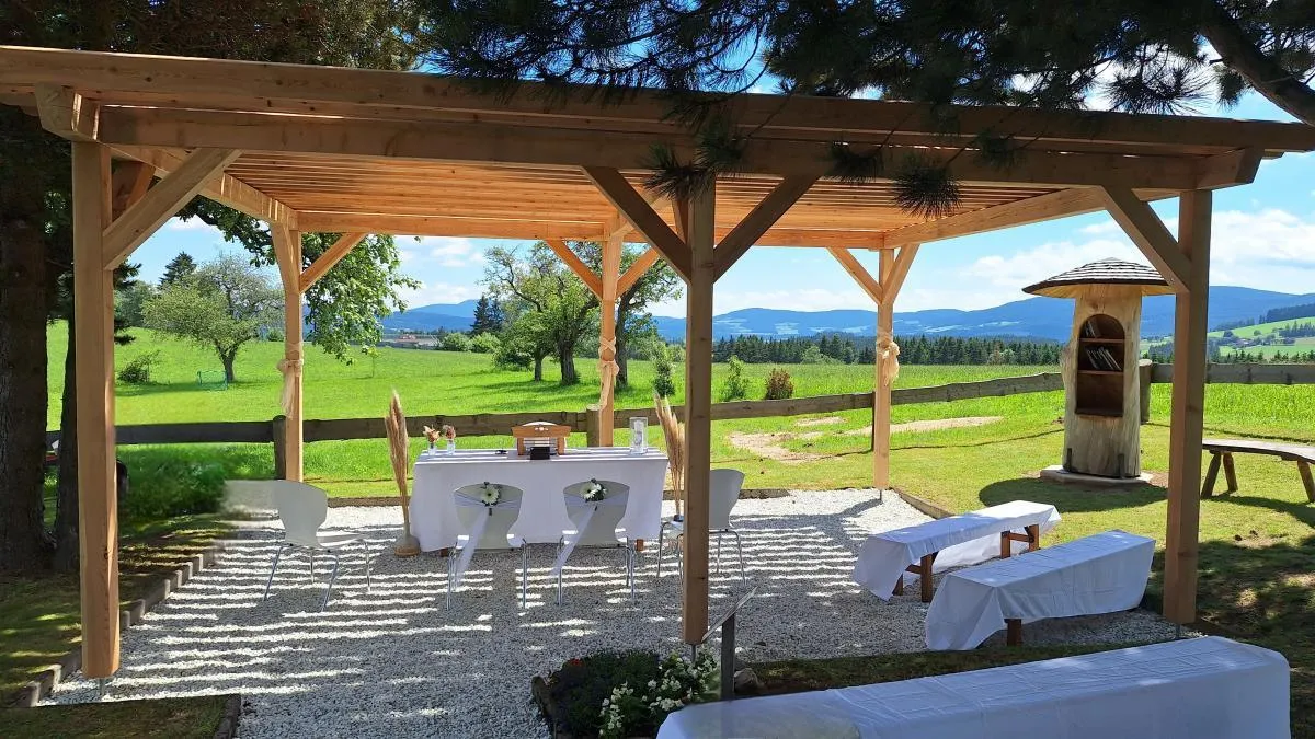 Eine offene Holzpergola mit einem gedeckten Tisch, umgeben von weißen Stühlen. Im Hintergrund befindet sich ein grasiger Bereich mit einem Zaun und Bäumen. In der Ferne sind Berge sichtbar.