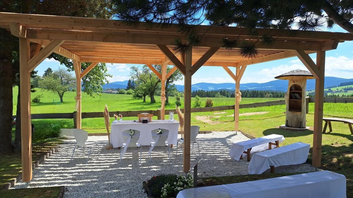 Eine offene Holzpergola mit einem gedeckten Tisch, umgeben von weißen Stühlen. Im Hintergrund befindet sich ein grasiger Bereich mit einem Zaun und Bäumen. In der Ferne sind Berge sichtbar.