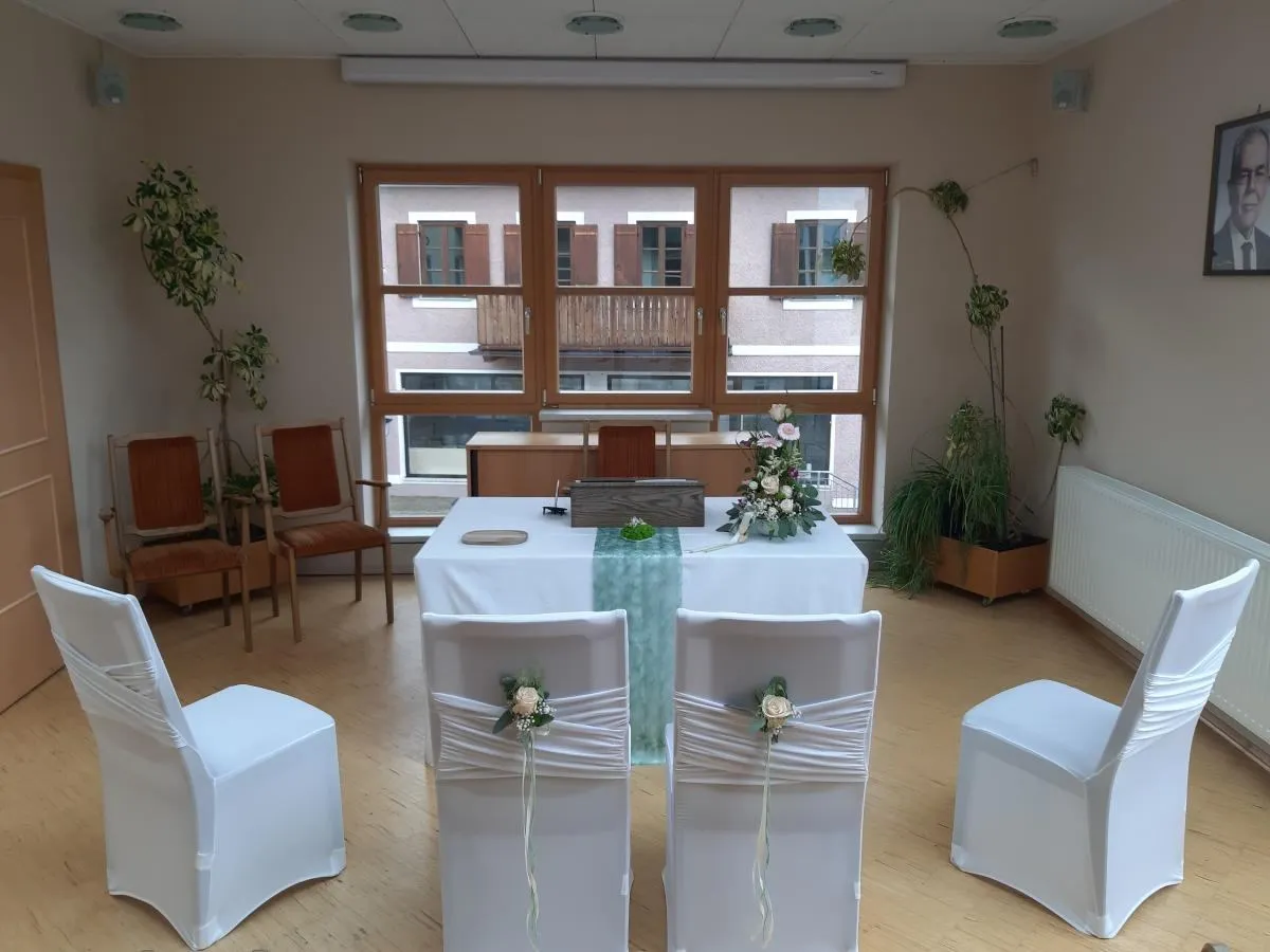 Ein Raum mit einem Tisch für eine Zeremonie, weißen Stühlen, Blumendekorationen und einem Fenster, das einen Balkon und ein Gebäude zeigt.