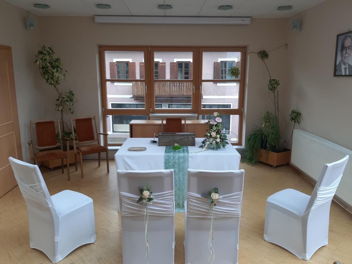 Ein Raum mit einem Tisch für eine Zeremonie, weißen Stühlen, Blumendekorationen und einem Fenster, das einen Balkon und ein Gebäude zeigt.