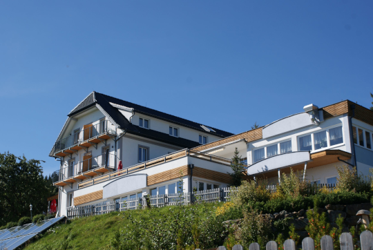 Ein großes weißes Gebäude mit schwarzem Dach und vielen Fenstern steht auf einem Hügel mit Grün und Solarpanelen.