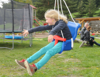 Ein junges Mädchen schwingt auf einem blauen und roten Schaukelstuhl und lächelt, während ein Erwachsener zusieht. Im Hintergrund sind ein Trampolin und ein hölzernes Spielhaus zu sehen.