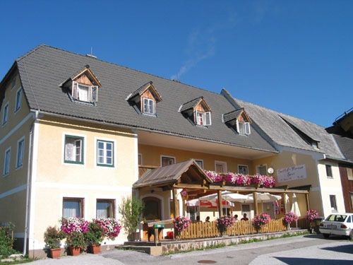 Ein gelbes Gebäude mit vielen Fenstern hat ein graues Dach und rosa Blumen. Es hat eine überdachte Veranda mit Tischen und Stühlen. Ein weißes Auto ist auf der Straße geparkt.