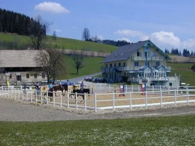 Ein Reitplatz mit Pferden und Reitern in einem eingezäunten Bereich. Ein großes gelbes Gebäude und ein kleineres Haus im Hintergrund.
