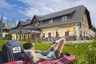 Eine Person ruht sich auf einer karierten Decke vor einem Gebäude mit dem Schild Orhhofen aus. Der Bereich ist von Gras umgeben und hat Schirme, Tische und Stühle.