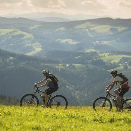 Zwei Radfahrer mit Helmen fahren auf einem Rasenfeld mit Fahrrädern. Sie tragen Rucksäcke. Dahinter ist das Land mit Bäumen und Hügeln bedeckt. Der Himmel ist klar mit ein paar Wolken.