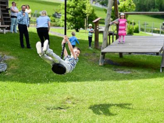 Ein Kind schwingt auf einem Reifenschaukel, während Erwachsene und Kinder in einem grünrasigen Außenbereich zuschauen.