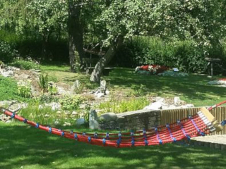Eine rote und blaue Hängematte hängt zwischen zwei Bäumen in einem Garten mit grünem Rasen und Bäumen.