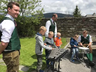 Eine Gruppe von Kindern und Erwachsenen spielt Instrumente auf einer Grasfläche. Zwei Kinder schlagen auf Trommeln, andere auf einem Xylophon. Ein Mann in traditioneller Kleidung beobachtet.