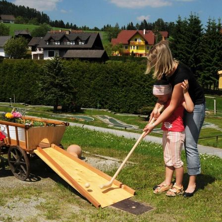 Eine Mutter hilft ihrer kleinen Tochter bei einem Minigolfspiel. Sie befinden sich auf einem Parcours, der von Häusern und Bäumen umgeben ist.