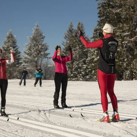 Eine Gruppe von Skifahrern befindet sich auf einer verschneiten Piste, gekleidet in Rot und Schwarz. Sie stehen und heben die Hände. Im Hintergrund sind zwei Personen beim Skifahren zu sehen. Es gibt mit Schnee bedeckte Tannenbäume.