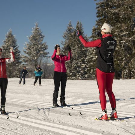Eine Gruppe von Skifahrern befindet sich auf einer verschneiten Piste, gekleidet in Rot und Schwarz. Sie stehen und heben die Hände. Im Hintergrund sind zwei Personen beim Skifahren zu sehen. Es gibt mit Schnee bedeckte Tannenbäume.