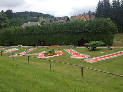 Ein Minigolfplatz liegt in einem grünen Feld mit einem Holzzaun auf der linken Seite. Der Platz hat rote Wege und der Rasen ist gut gepflegt. Hinter dem Hecken befinden sich Häuser und Bäume. Der Himmel ist bewölkt.