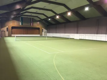 Ein überdachter Tennisplatz mit neuem grünem Belag. Der Platz hat weiße Linien und ein Tennisnetz in der Mitte. Die Decke hat mehrere Lichter.