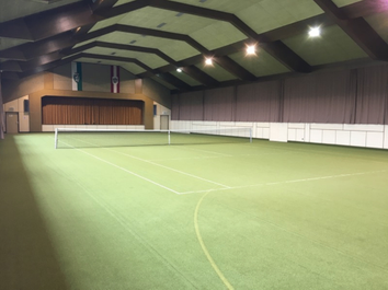 Ein überdachter Tennisplatz mit neuem grünem Belag. Der Platz hat weiße Linien und ein Tennisnetz in der Mitte. Die Decke hat mehrere Lichter.