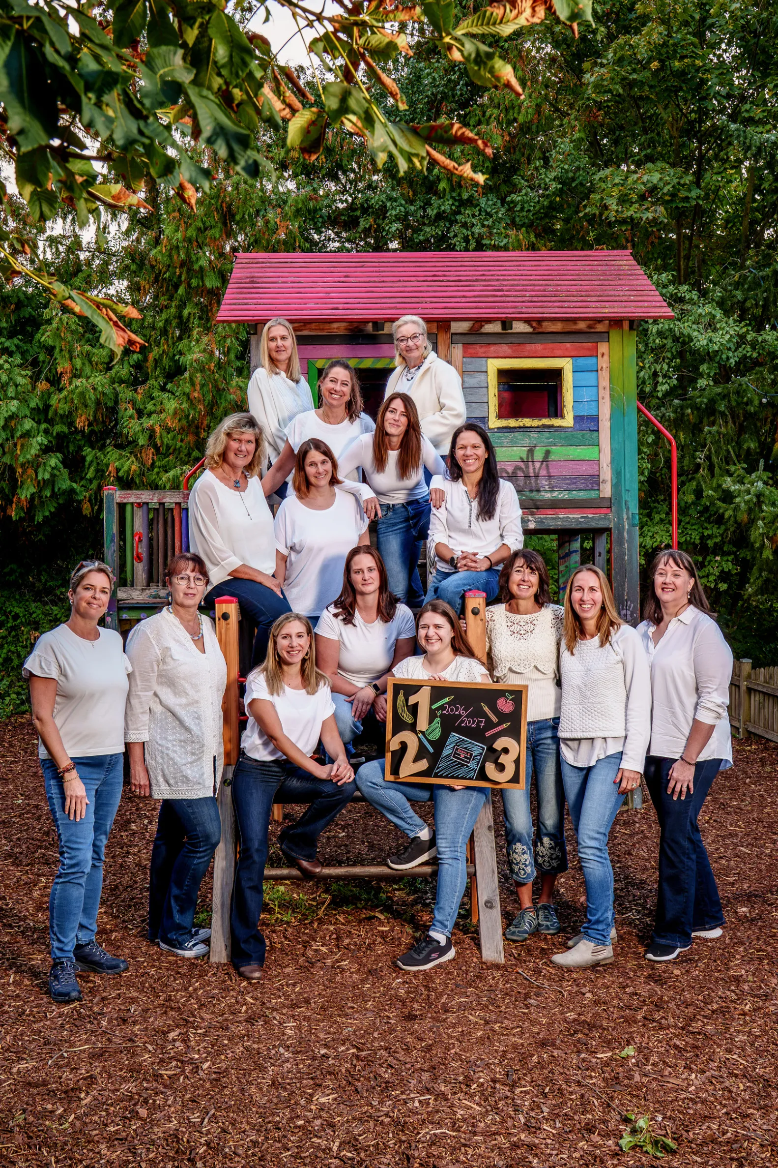 Eine Gruppe von Frauen in weißen Shirts und Jeans posiert für ein Foto vor einem bunten Spielhaus in einem Park.