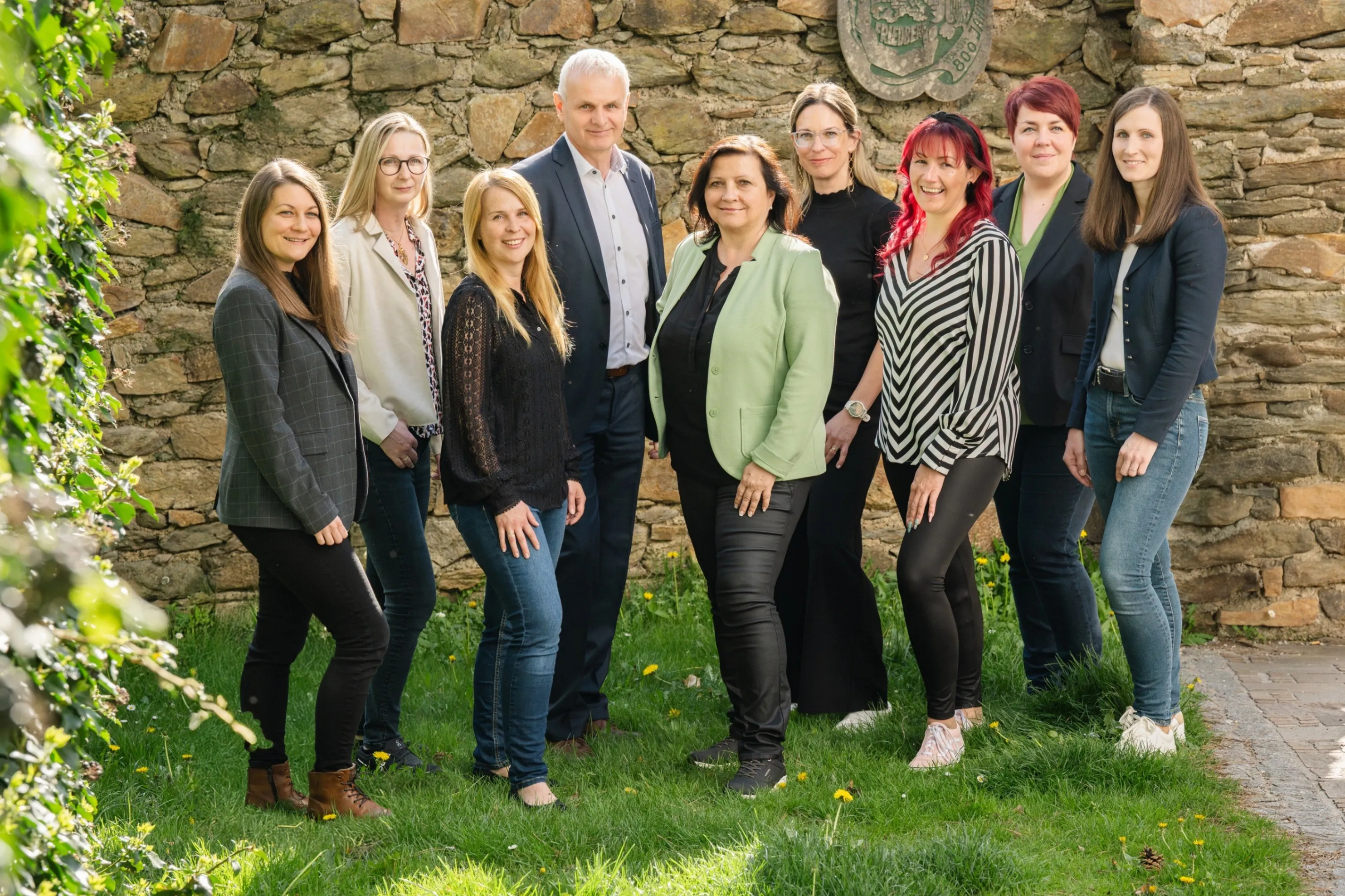 Eine Gruppe lächelnder Frauen und Männer, möglicherweise ein Team, steht vor einer Steinmauer.