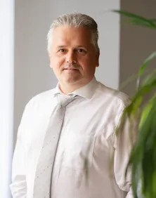 Ein Mann mit weißen Haaren steht neben einer grünen Pflanze, trägt ein weißes Hemd und eine graue Krawatte. Er lächelt und schaut nach vorne.