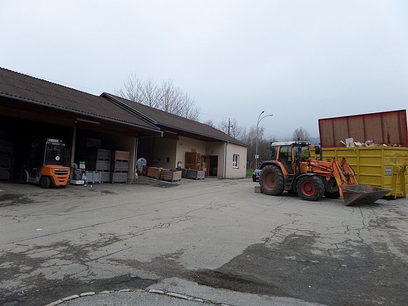 Ein orangefarbener Gabelstapler ist in einem Lagerhaus unter einem braunen Dach geparkt. Vor dem Lagerhaus steht ein gelber Müllcontainer, und ein großer orangefarbener Traktor mit Schaufel ist rechts geparkt.