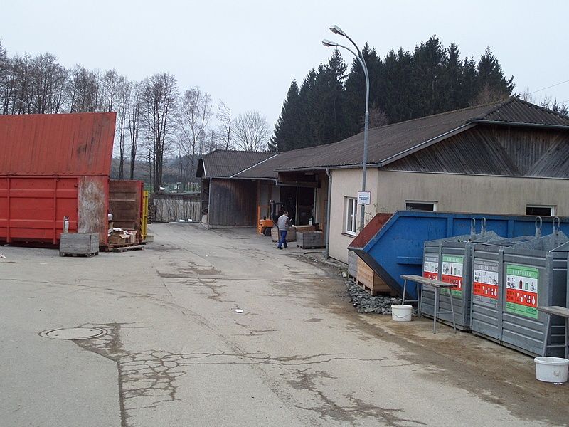 Eine Straße mit einem Gebäude rechts, einem blauen Müllcontainer, einem roten Müllcontainer und zwei Personen vor dem Gebäude.