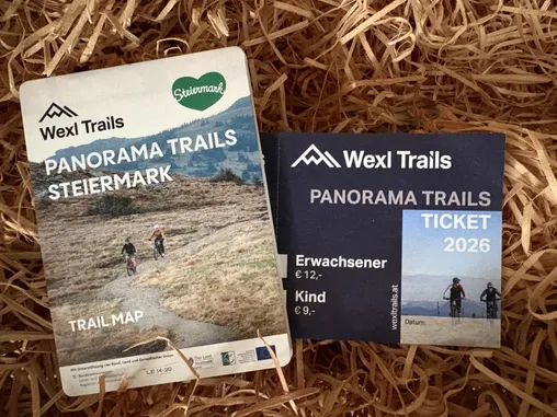 Eine Sammlung von Broschüren und Tickets für die Wexl Trails, einschließlich einer Streckenkarte und Preisen für Erwachsene und Kinder. Die Broschüren haben unterschiedliche Farben und liegen auf einem Strohbett.