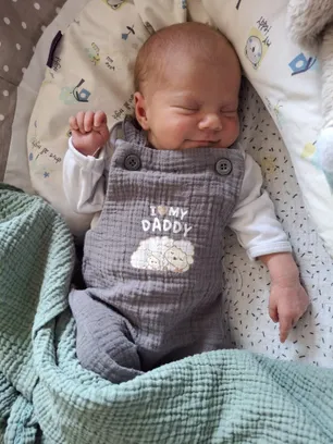 Ein Baby schläft auf einem Bett mit einer Decke und einem Kissen. Das Baby trägt einen grauen Overall mit den Worten 'Ich liebe meinen Daddy'.