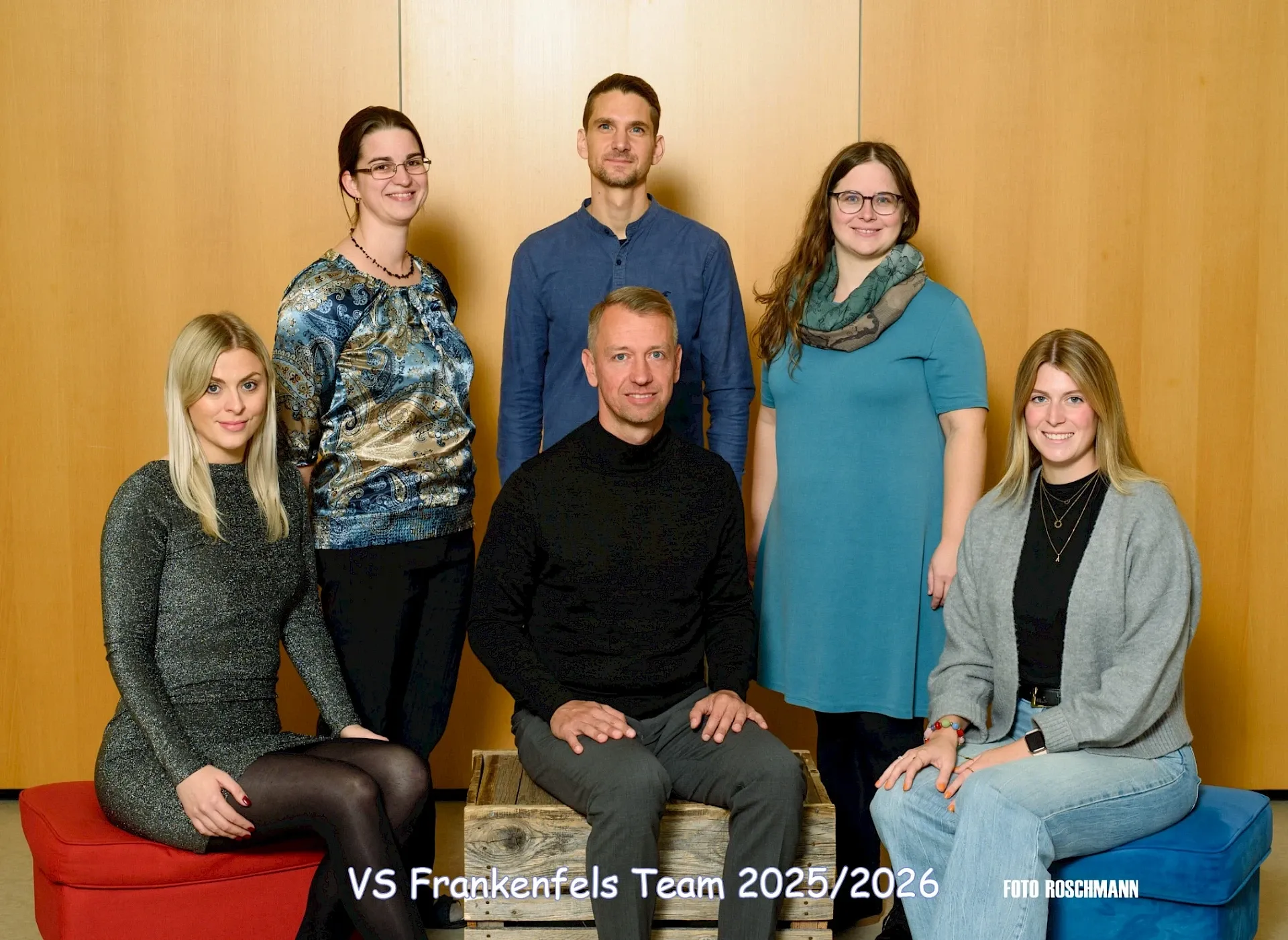Eine Gruppe von sechs Personen, vier Frauen und zwei Männer, posiert für ein Foto. Sie stehen und sitzen auf Holzkisten, wobei ein Mann in der Mitte sitzt. Sie alle lächeln und scheinen für ein Teamfoto zu posieren.