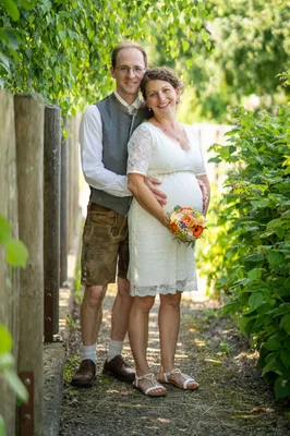 Ein Mann und eine schwangere Frau stehen lächelnd in einem Garten und halten einen Blumenstrauß. Der Mann trägt eine Weste und Shorts, die Frau ein weißes Kleid.