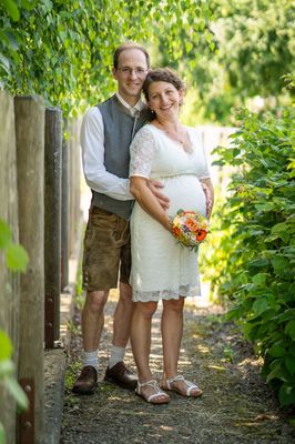 Ein Mann und eine schwangere Frau stehen lächelnd in einem Garten und halten einen Blumenstrauß. Der Mann trägt eine Weste und Shorts, die Frau ein weißes Kleid.