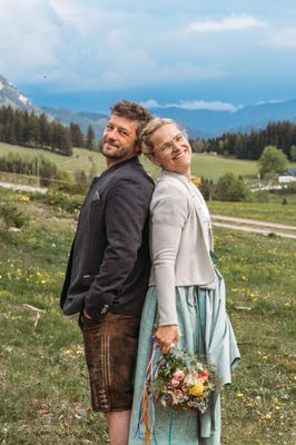 Ein Paar steht in einem Grasfeld mit Bergen im Hintergrund, beide lächeln und halten Blumen.