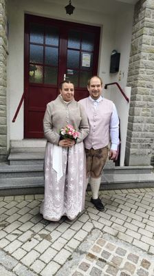 Ein Mann und eine Frau in traditioneller Kleidung stehen vor einem Gebäude mit Ziegelboden. Die Frau hält einen Blumenstrauß.