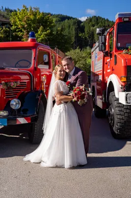Ein frisch verheiratetes Paar steht vor zwei roten Feuerwehrautos, die Braut in einem weißen Kleid und der Bräutigam im Anzug, beide lächeln.