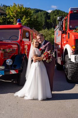 Ein frisch verheiratetes Paar steht vor zwei roten Feuerwehrautos, die Braut in einem weißen Kleid und der Bräutigam im Anzug, beide lächeln.