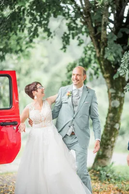 Eine lächelnde Braut in einem weißen Kleid und ein Bräutigam in einem hellblauen Anzug stehen vor einem roten Auto, mit einem Baum und Grün im Hintergrund.