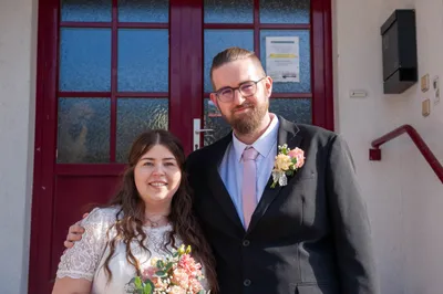 Ein Mann und eine Frau stehen lächelnd vor einer roten Tür. Der Mann trägt einen schwarzen Anzug und eine pinke Krawatte mit einer Blume auf der Brust. Die Frau hält einen Blumenstrauß und trägt ein weißes Kleid.