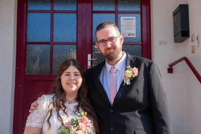 Ein Mann und eine Frau stehen lächelnd vor einer roten Tür. Der Mann trägt einen schwarzen Anzug und eine pinke Krawatte mit einer Blume auf der Brust. Die Frau hält einen Blumenstrauß und trägt ein weißes Kleid.