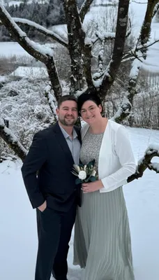 Ein Mann und eine Frau stehen eng beieinander in einer verschneiten Landschaft, lächeln und posieren für ein Foto. Der Mann trägt einen dunklen Anzug, und die Frau hält einen Blumenstrauß. Sie stehen unter einem mit Schnee bedeckten Baum.