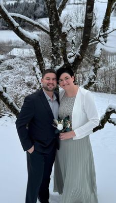 Ein Mann und eine Frau stehen eng beieinander in einer verschneiten Landschaft, lächeln und posieren für ein Foto. Der Mann trägt einen dunklen Anzug, und die Frau hält einen Blumenstrauß. Sie stehen unter einem mit Schnee bedeckten Baum.