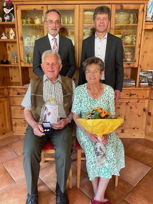 Ein älteres Paar, beide lächelnd, sitzt in einem Raum mit Holzschränken. Der Mann hält ein Geschenk, und die Frau hält einen Blumenstrauß. Zwei Männer stehen hinter ihnen, einer trägt eine Krawatte.