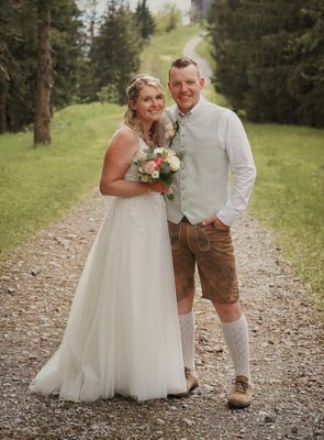 Ein frisch verheiratetes Paar steht auf einem Kiesweg, die Braut in einem weißen Kleid und der Bräutigam in traditioneller Kleidung, beide lächeln.