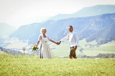 Ein Paar steht Hand in Hand auf einem Grashügel, mit Bergen und einem Tal im Hintergrund. Die Frau hält einen Blumenstrauß.