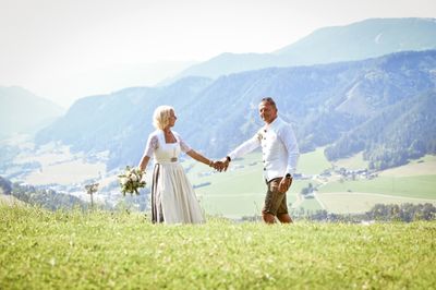 Ein Paar steht Hand in Hand auf einem Grashügel, mit Bergen und einem Tal im Hintergrund. Die Frau hält einen Blumenstrauß.