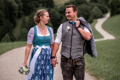 Ein lächelndes Paar in traditioneller deutscher Kleidung, Hand in Hand auf einem Weg in einem Park.