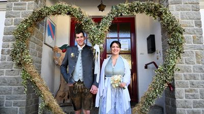 Ein frisch verheiratetes Paar steht vor einem geschmückten Bogen und lächelt in die Kamera. Der Mann trägt ein traditionelles deutsches Outfit und die Frau hält einen Blumenstrauß. Hinter ihnen befindet sich eine rote Tür mit einem Fenster.