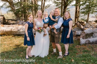 Eine Familie mit fünf Personen steht in einer Waldlichtung, wobei die Braut in einem weißen Kleid, der Bräutigam in traditioneller Kleidung und zwei kleine Kinder in ihren Armen sind.